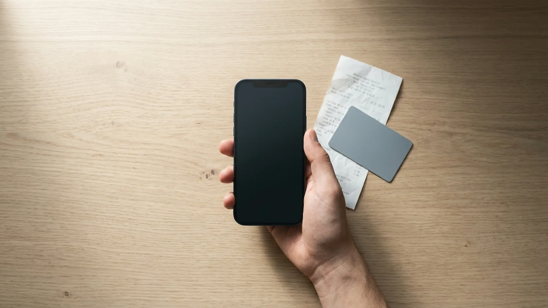 Close-up of a phone held over a wooden desk showing a payment confirmation, with a bank card and a paper receipt beside it in soft window light
